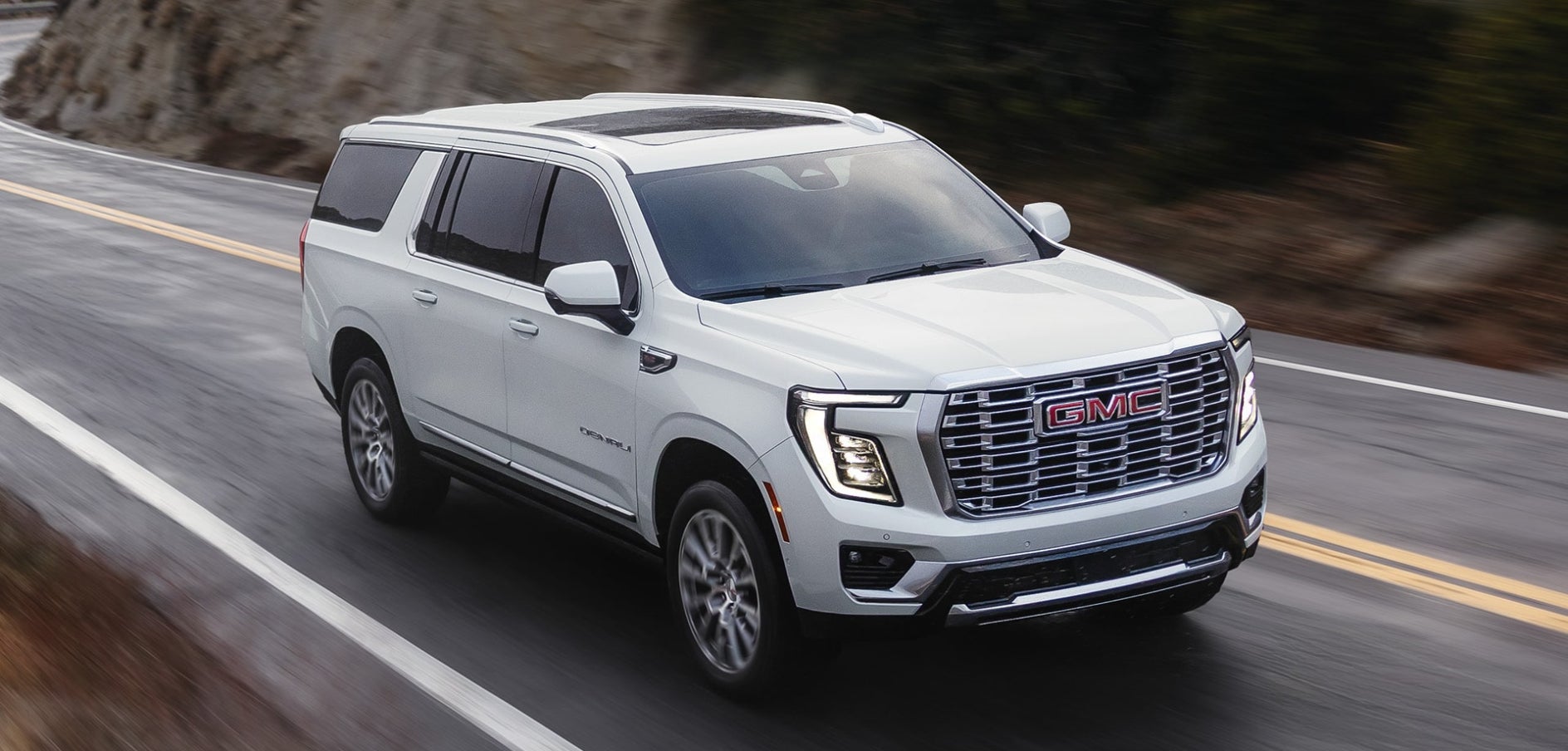 A white GMC Yukon driving down the road by a cliffside.